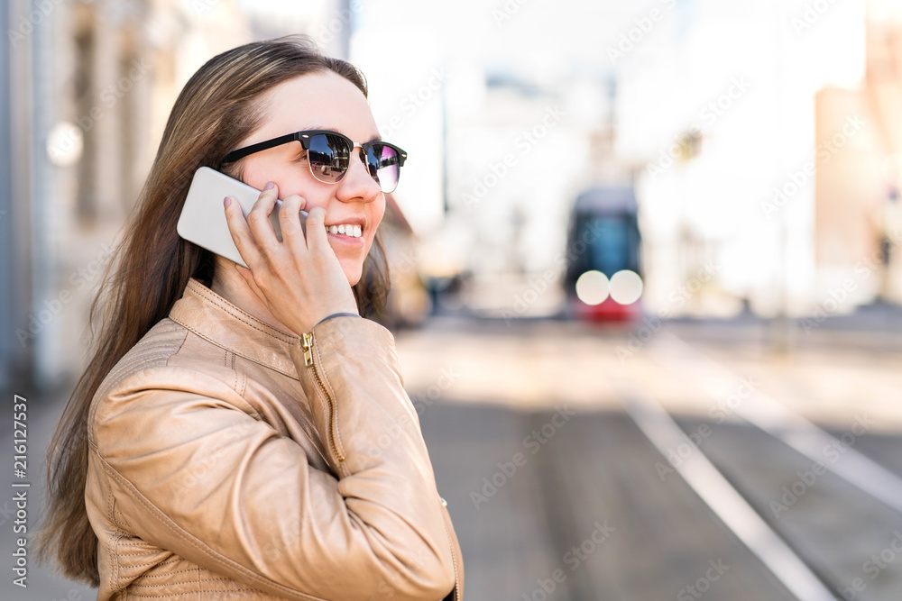 Young woman waiting for city train and talking on the phone. Passenger at tram stop calling with smartphone. Public transport and communication concept.