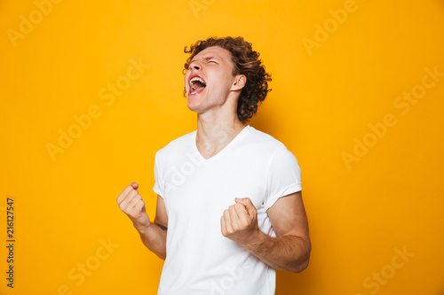 Photo of happy man 20s in casual t-shirt rejoicing and screaming with clenching fists, isolated over yellow background photo