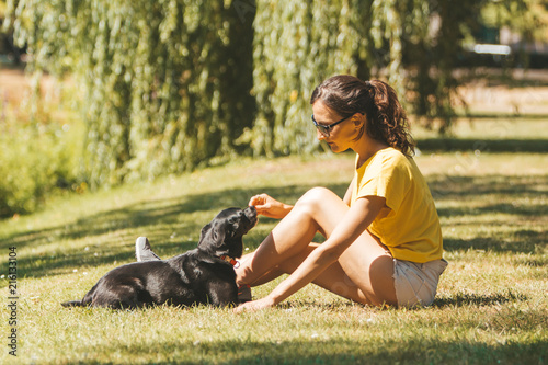 A young woman is sitting on the grass and playing (or feeding) with a dog / picture of a girl with sporty outfit having fun in a sunny garden