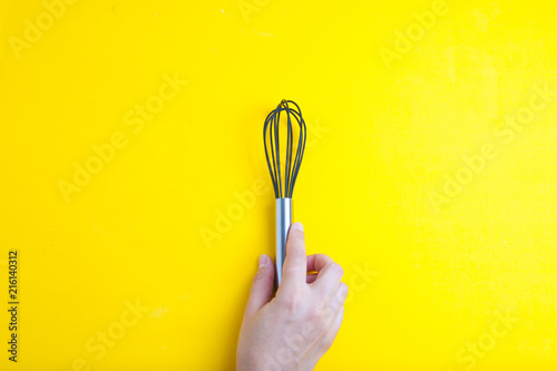 Female hand with a kitchen whisk onyellow background isolated with copy space for text. Top view. photo