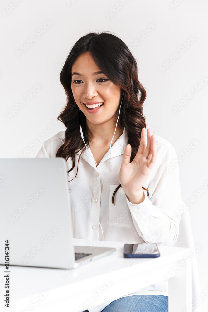 Image of young asian business woman 20s wearing office clothing and earphones waving at laptop, during video call or online conference, isolated over white background Image of young asian business woman 20s wearing office clothing and earphones waving at laptop, during video call or online conference, isolated over white background