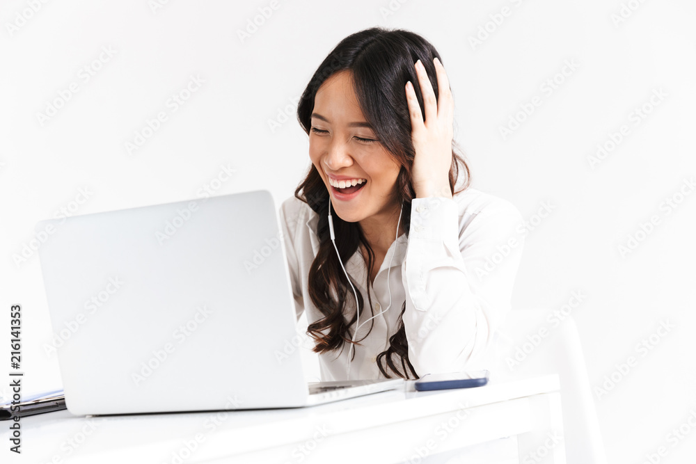 Attractive asian woman 20s wearing office clothing and earphones looking at laptop and laughing, during video call or online conference, isolated over white background Attractive asian woman 20s wearing office clothing and earphones looking at laptop and laughing, during video call or online conference, isolated over white background