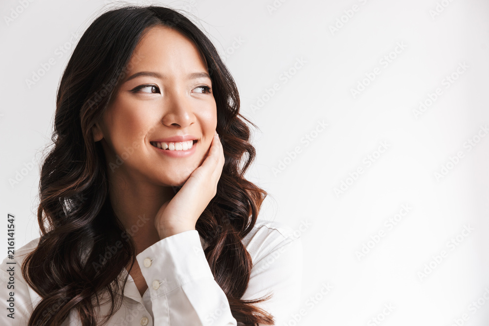 Naklejka premium Image of smiling chinese woman with long dark hair looking aside at copyspace and touching cheek, isolated over white background in studio