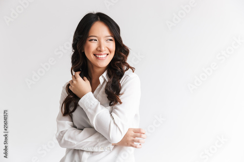 Photo of optimistic asian woman with long dark hair looking aside at copyspace and laughing, isolated over white background in studio