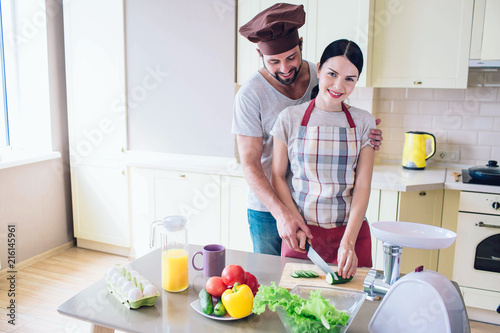 Beautiful couple stands at kitchen and wokr together. Girl looks at camera and smiles. She holds knife in one hand and cucumber in the other one. Guy helps her to cut vegetable.