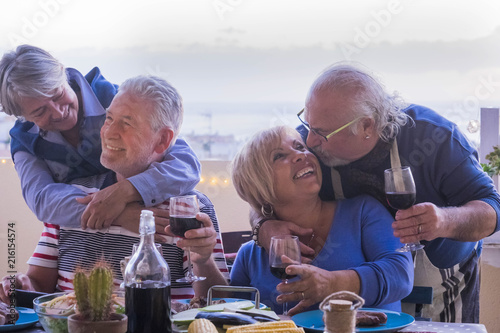 causasian mature couples friends having fun during evening dinner together. kiss and hug and smile and laugh for great retired lifestyle concept. outdoor on the terrace with ocean view. 
