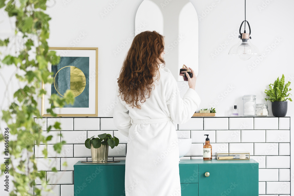 Φωτογραφία Woman in front of mirror in white bathroom interior with poster, plants and green cabinet