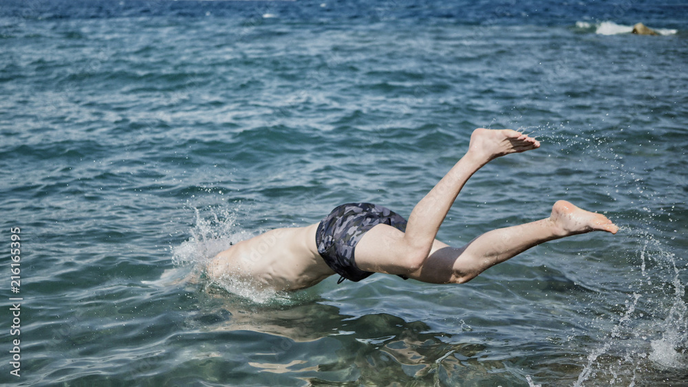 Attractive young shirtless athletic man jumping in water by sea or ocean shore, wearing shorts