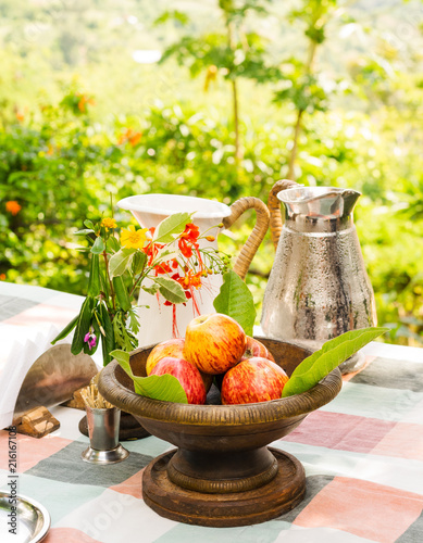 A copper bowl of red apples and pitchers on a checkered table cloth, overlooking a landscape. photo