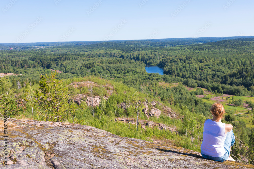 The green forest of fir, spruce an pine trees near the shore of the Ladoga in Russia lake in the sunny summer day and the girl sitting on the stone