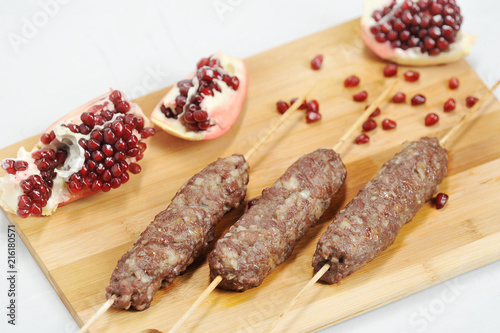 Lulya kebab on a wooden board. Next to the kebab is a pomegranate grain and a pomegranate fruit, broken into several pieces. White background. Close-up. View from above.