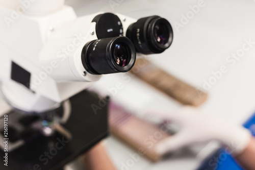The eyepieces of the microscope are close-up. In the background, the lab technician's hands take samples.