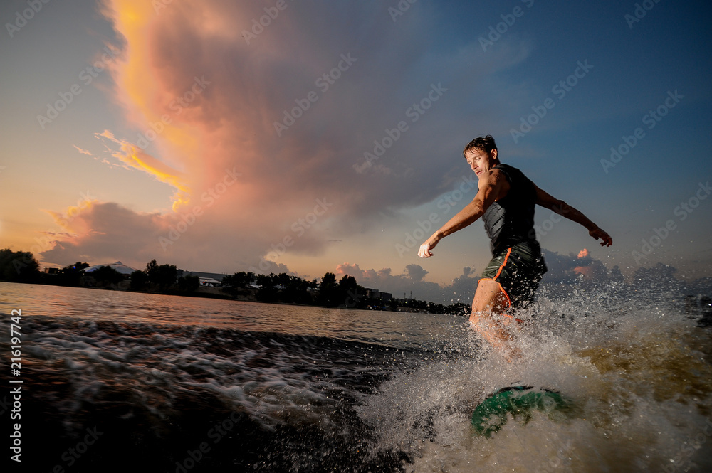 Young and athletic man riding on wakesurf down the river during sunset