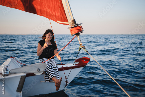 Young pretty woman in black shirt and striped skirt with a glass at luxury yacht in sea, sunset