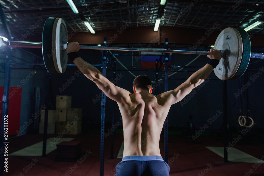 Portrait of a handsome athlete from behind. Athlete raises the barbell over your head. Studio shots in the dark tone. Cross style fit, deadlift Portrait of a handsome athlete from behind. Athlete raises the barbell over your head. Studio shots in the dark tone. Cross style fit, deadlift