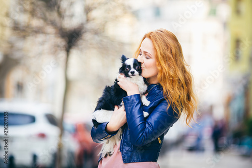 A young redhaired Caucasian woman with freckles holds and kisses, embracing black and white shaggy dog of Chihuahua breed. girl dressed in blue leather jacket, stands on street in spring in Europe photo