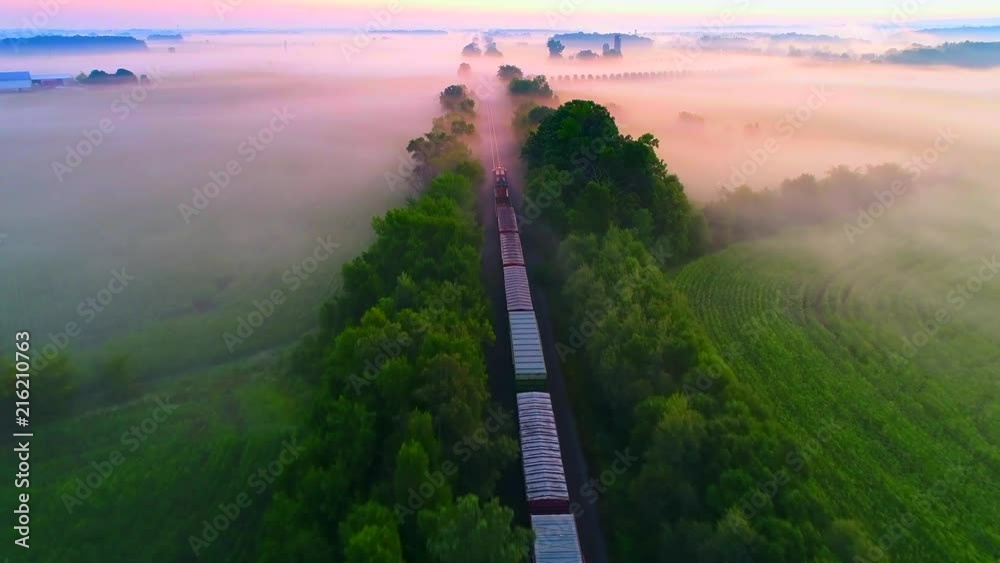 Freight train rolls across foggy rural landscape at sunrise with ethereal beauty, aerial view.