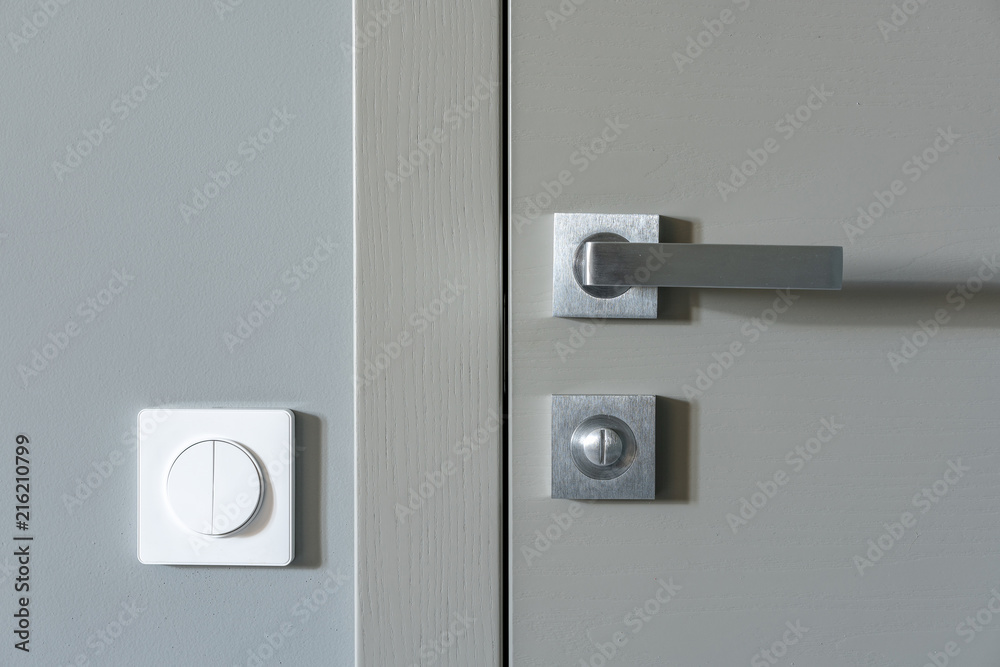 A metal handle and a lock on the gray interior door and a white switch on the gray wall. Element of apartment design. A metal handle and a lock on the gray interior door and a white switch on the gray wall. Element of apartment design.