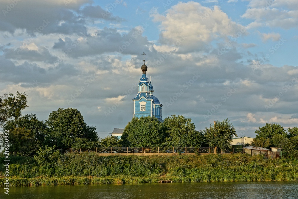 blue old wooden church in green vegetation on the shore of the reservoir against the sky and clouds