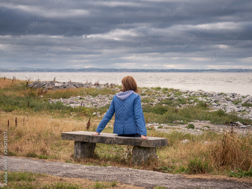 Woman sitting on a bench looking out landscape around her, ocean, cloudy sky. 