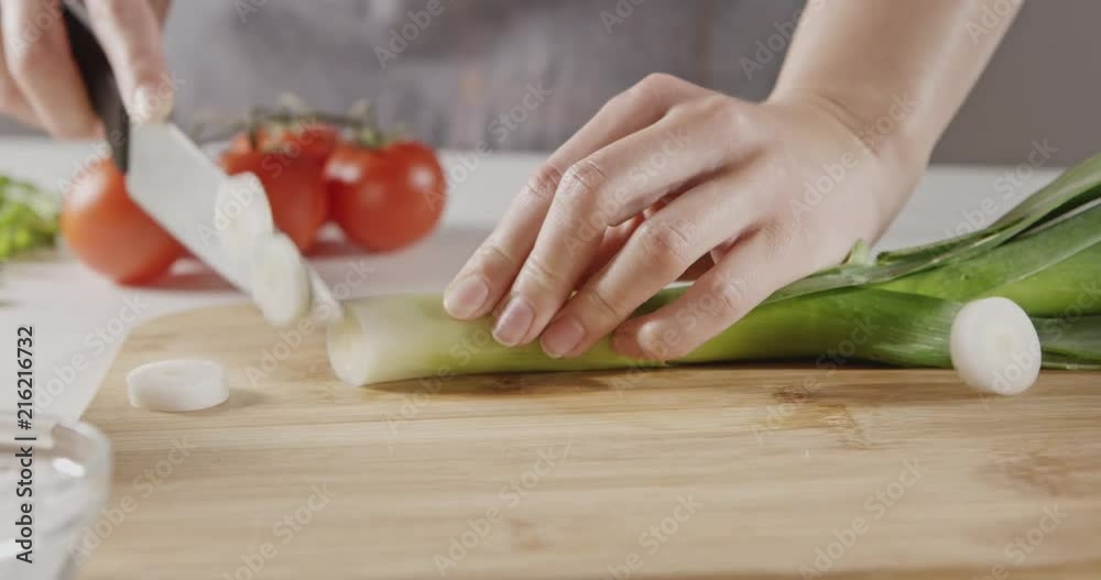A woman in an apron slices green leeks on a wooden cutting board for salad preparation on a white kitchen table with red tomatoes, greens. Close-up. Slow motion video in 4K.
