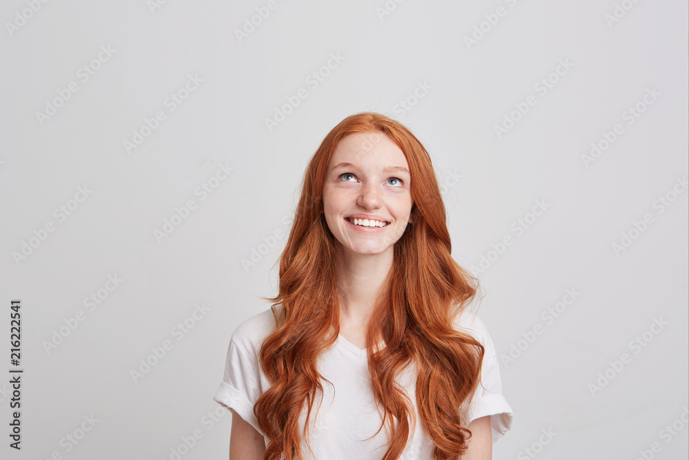 Closeup of cheerful beautiful young woman with long wavy red hair and freckles wears t shirt feels happy and looks up isolated over white background Closeup of cheerful beautiful young woman with long wavy red hair and freckles wears t shirt feels happy and looks up isolated over white background