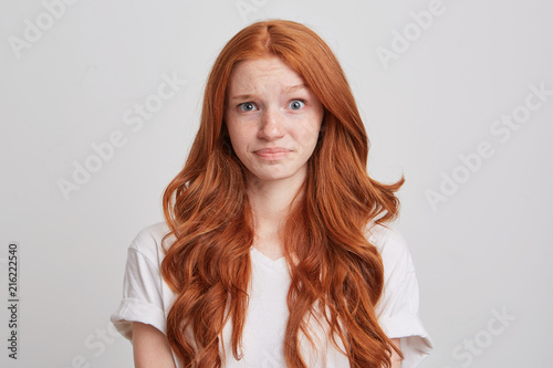 Portrait of confused astonished young woman with long wavy red hair and freckles wears t shirt feels embarrassed and looks directly in camera isolated over white background
