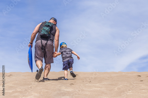 Father and son walking up hand in hand to go sand sliding in dessert with a snow sled