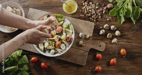 A woman's hand sprinkles with spices salad from natural ingredients. Top view of background with quail eggs, chicken fillet, greens, tomatoes, walnuts for cooking salad. Slow motion video in 4K