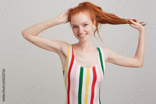 Portrait of happy beautiful young woman with red hair and freckles in striped top holding her ponytail and posing isolated over white background Looks satisfied