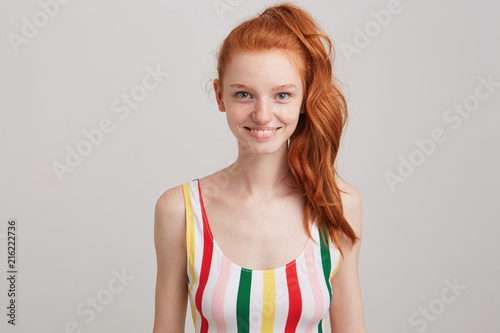 Closeup of smiling attractive redhead young woman with freckles and ponytail wears striped top feels confident and looks directly in camera isolated over white wall