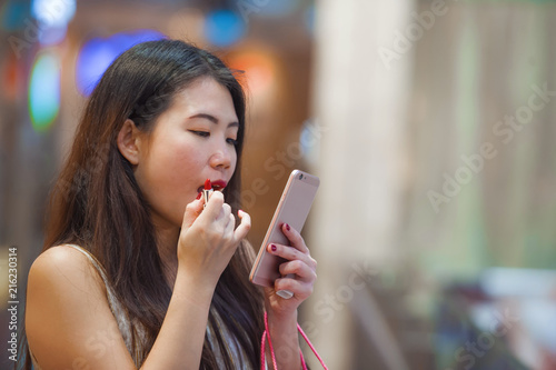 beautiful and happy Asian Chinese woman retouching lips with lipstick makeup looking at mobile phone using it as mirror carrying shopping bags in fashion mall