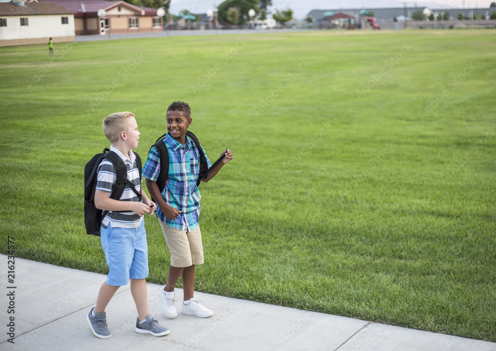 Two diverse school kids walking home together after school and talking together. Back to school photo of diverse school children wearing backpacks in the school yard Two diverse school kids walking home together after school and talking together. Back to school photo of diverse school children wearing backpacks in the school yard