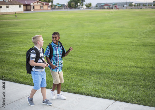 Two diverse school kids walking home together after school and talking together. Back to school photo of diverse school children wearing backpacks in the school yard