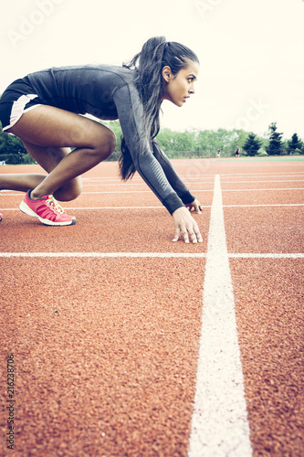 Athlete crouched in starting position on a running track. Ethnic, multicultural theme. Wearing long sleeve black top, shorts and pink runners. Long black hair in a pony tail.