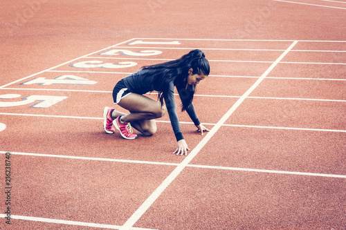 Woman crouched in starting position on a running track. Ethnic, multicultural theme. Wearing long sleeve black top, shorts and pink runners. Long black hair in a pony tail.