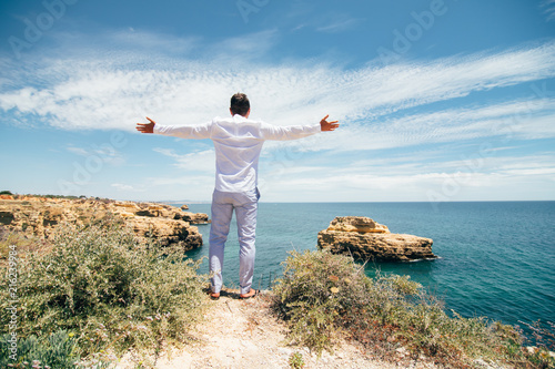 Young handsome man with raised hands at the edge of cliff enjoy the freedom of ocean view. Freedom concept. © F8  \ Suport Ukraine
