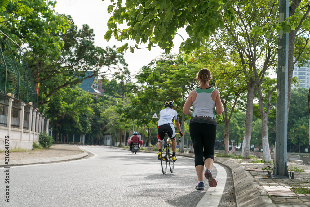 Sporty Caucasian woman running on the road in the Asian city park with green tree row in early morning