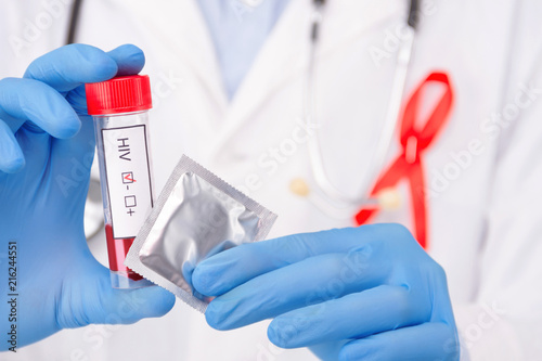 Safe sex / healthcare concept. Doctor wearing white coat with pinned red ribbon as a symbol of hiv aids and stethoscope holding negative tumor marker / blood test and condom. 