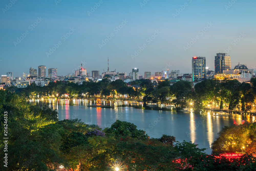 Fototapeta premium Hoan Kiem lake lub Sword lake, Ho Guom w Hanoi, Wietnam z Turtle Tower, zielone drzewa i budynki na horyzoncie, o zmierzchu