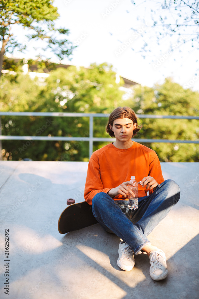 Obraz premium Young guy in orange pullover with skateboard holding bottle of water in hand while spending time at skatepark