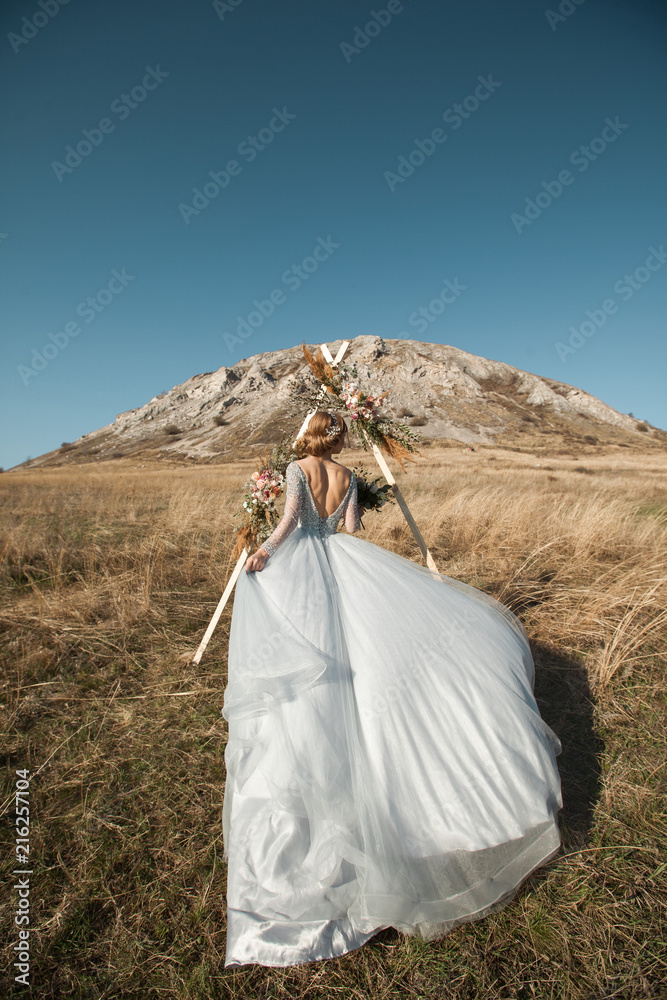 beautiful young girl with make-up and hairstyle standing in summer in a field with a bouquet on the background of a mountain