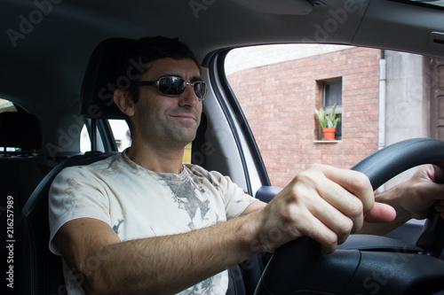 Shot from inside car of happy man driving without seatbelt on in urban city. Smiling young driver with sunglasses and white shirt. Road trip, auto rental, insurance traffic violation concepts