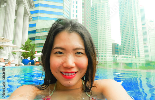  young happy and beautiful Asian Korean tourist woman taking selfie picture with mobile phone camera at luxury hotel infinity pool with modern buildings urban background © TheVisualsYouNeed