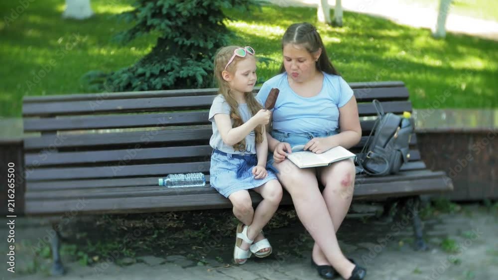 Two little girls are sitting on a wooden bench in a city reading a book and eating ice cream, the background of a city park Two little girls are sitting on a wooden bench in a city reading a book and eating ice cream, the background of a city park