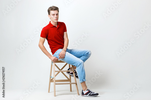 Calm handsome dark-haired guy wearing red shirt and blue jeans sitting on wooden stool over white background