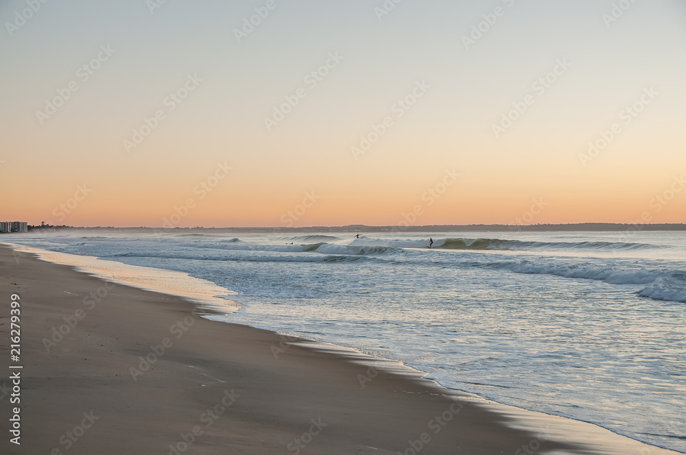 A gentle early morning on the shore of the Atlantic Ocean. Surfers riding on waves in the distance. USA. Maine.
