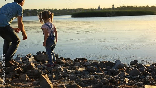 bearded father with a little girl sitting by the river and throw stones into the water on a summer evening at sunset. photo