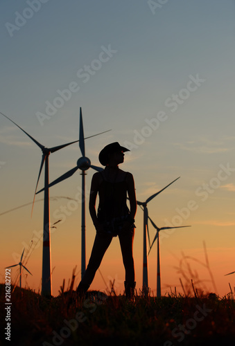 A young girl stands in a field and admires the wind turbines in the distance. The sun is setting and the sunlight creates a silhouette to the scenery. 