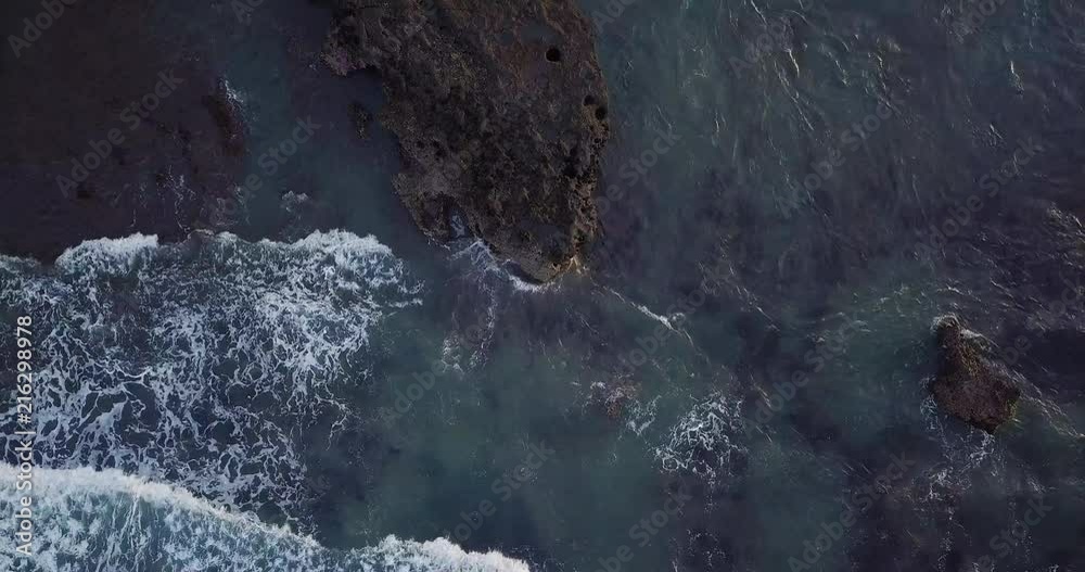 Aerial top view waves break on dark rocks near beach. Sea waves on the dangerous stones aerial view drone 4k shot. Bird's eye view of ocean waves crashing against an empty beach from above. Aerial top view waves break on dark rocks near beach. Sea waves on the dangerous stones aerial view drone 4k shot. Bird's eye view of ocean waves crashing against an empty beach from above.